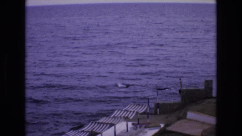 1972 Spain 8mm frame looking across a coastal terrace with striped beach shelters, stone walls, white buildings, and the purple-tinted sea beyond.