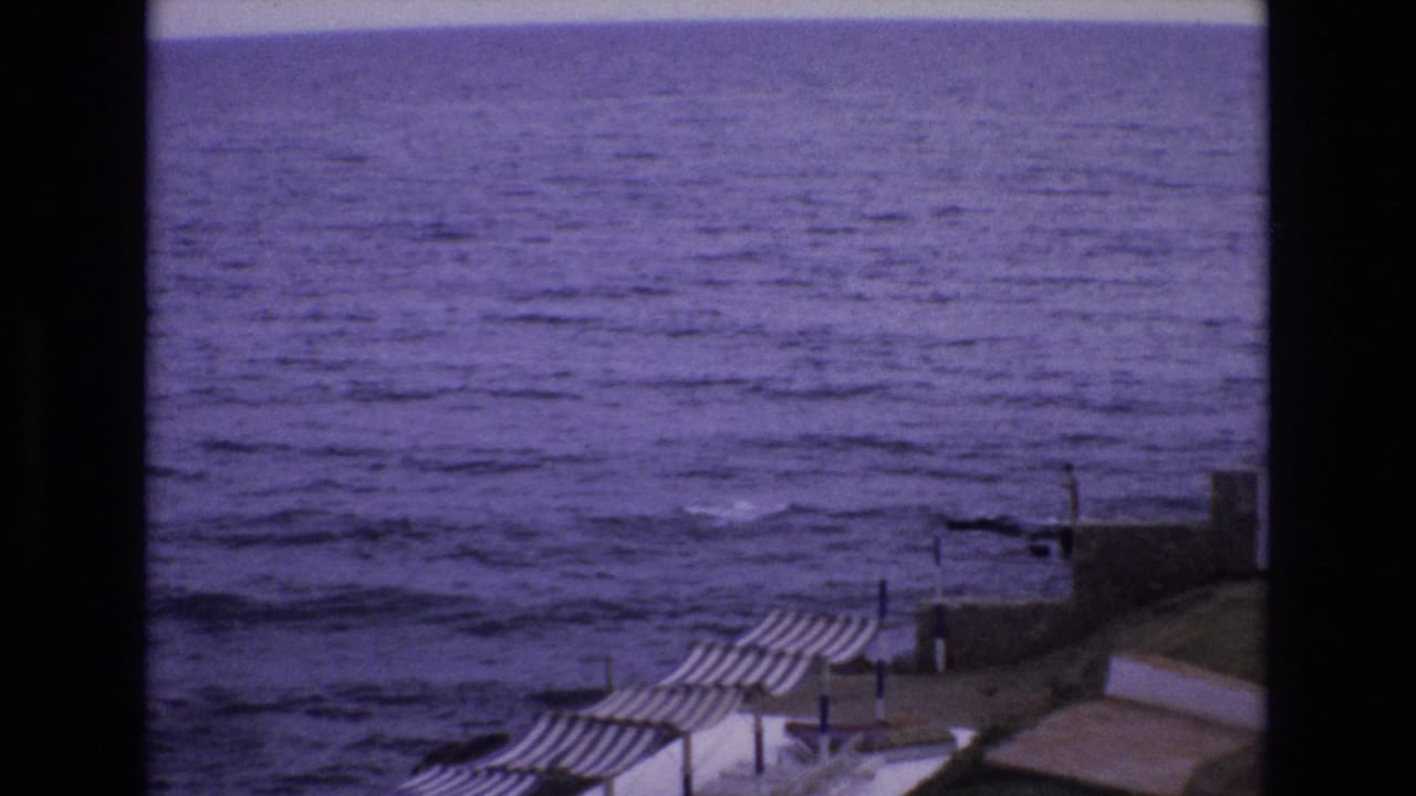 Elevated 8mm view of the ocean and striped beach umbrellas along a Spanish seaside promenade, dated 1972 by clip metadata.