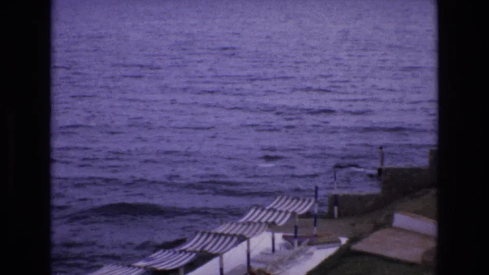 1972 Spain home-movie frame showing blue ocean water, a seawall, and striped umbrellas arranged along the waterfront.