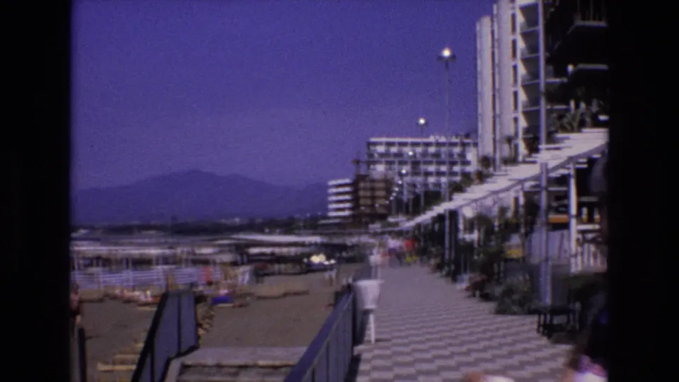 1972 Spain home-movie view of a seaside promenade lined with resort buildings, with distant mountains and small pedestrians visible.
