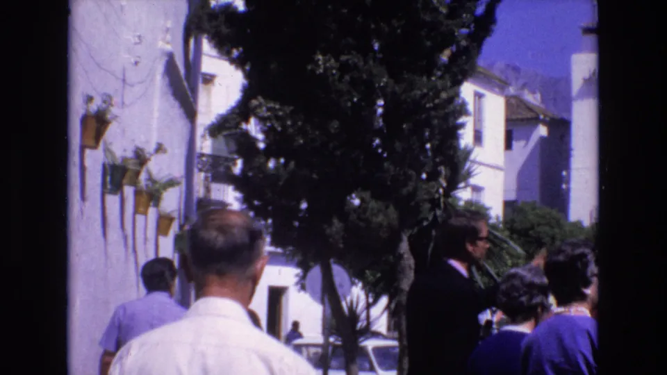 1972 Spain home-movie frame showing pedestrians in period clothing walking on a sunny narrow street with white buildings, trees, and parked cars in the distance.