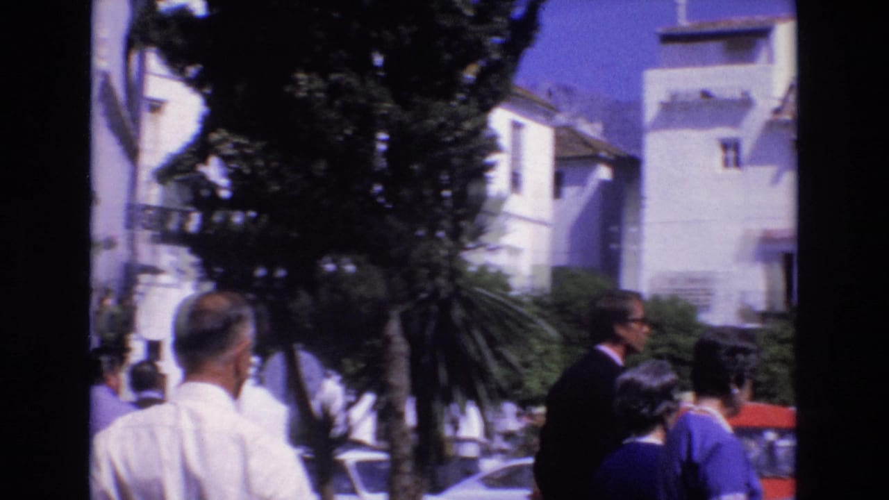 1972 Spain home-movie frame showing pedestrians near whitewashed buildings, trees, parked cars, and a sunlit public square.