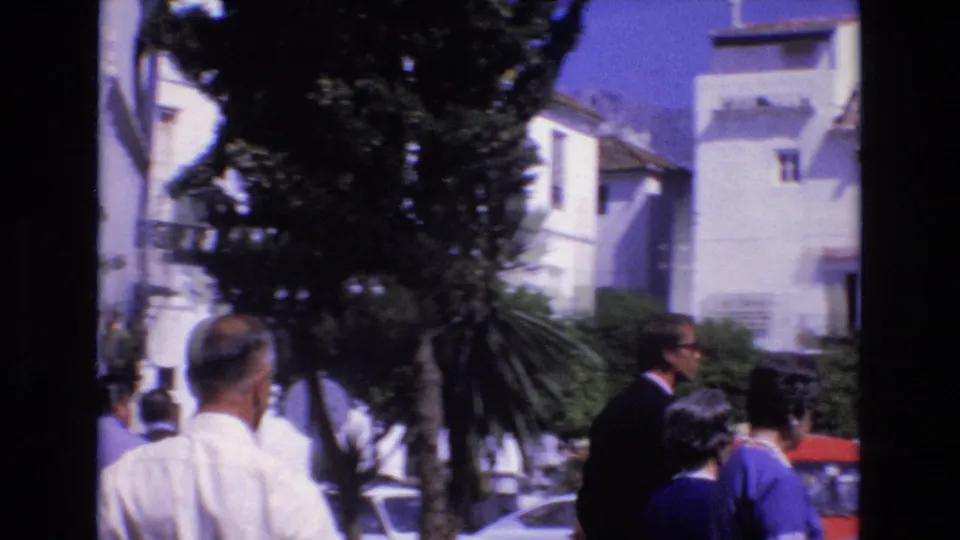 1972 Spain home-movie frame showing pedestrians near whitewashed buildings, trees, parked cars, and a sunlit public square.