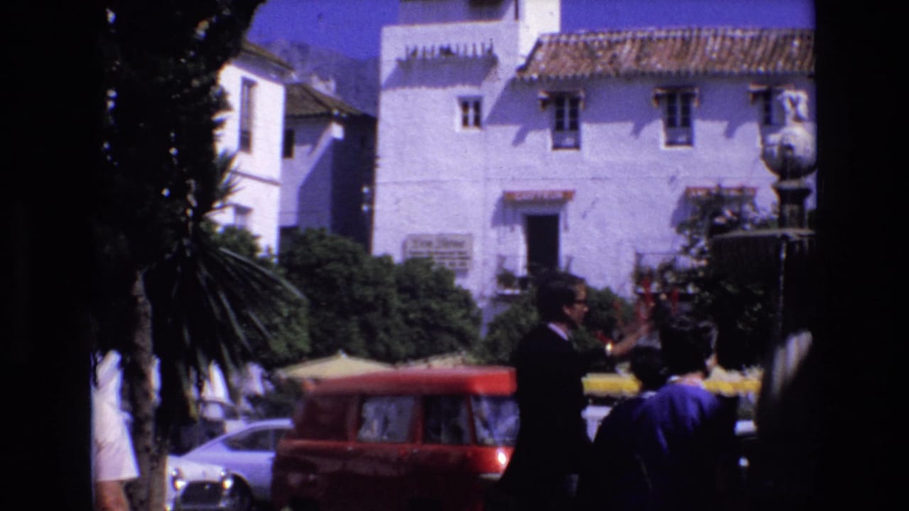 People stand near a fountain, red van, parked cars, and white buildings.