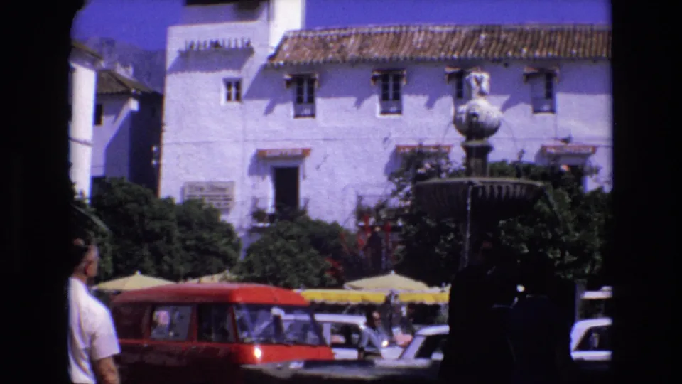 1972 Spain frame looking across a public square toward a whitewashed building, fountain, yellow awnings, red van, and pedestrians at the edge of the shot.