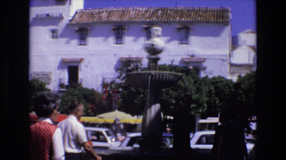 1972 Spain home-movie frame with pedestrians crossing the foreground, parked cars, a fountain, shrubs, and a white building with terracotta roof tiles.