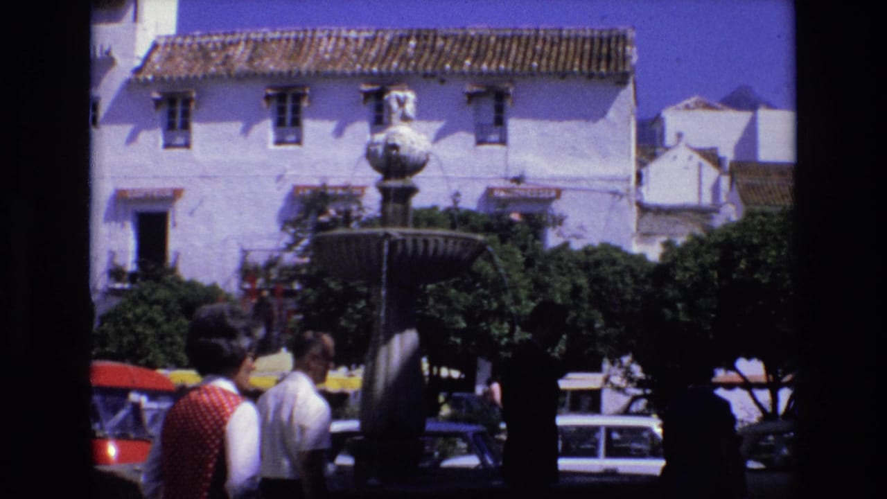 1972 Spain street scene showing pedestrians near a stone fountain, whitewashed buildings, terracotta roofs, trees, and parked compact cars in bright sun.