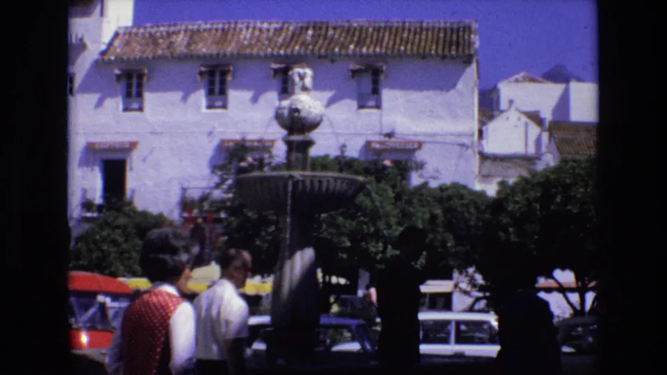 1972 Spain street scene showing pedestrians near a stone fountain, whitewashed buildings, terracotta roofs, trees, and parked compact cars in bright sun.