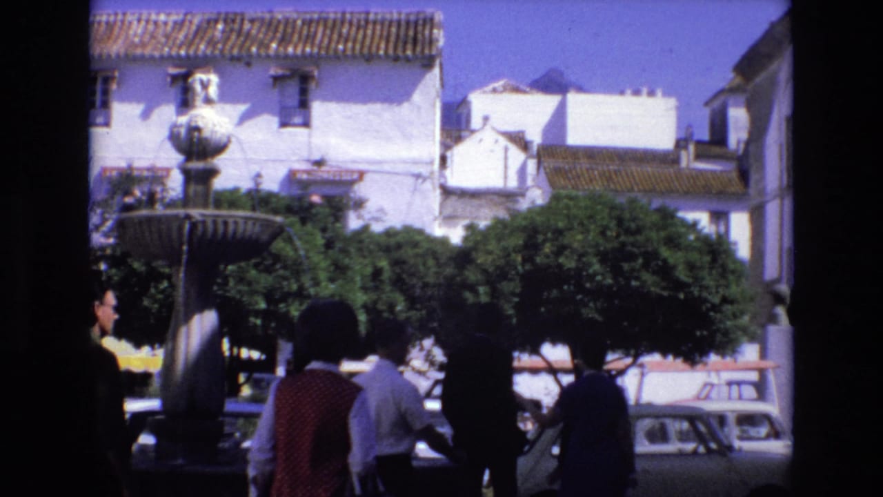A fountain stands in front of white buildings as pedestrians cross the square near parked cars.