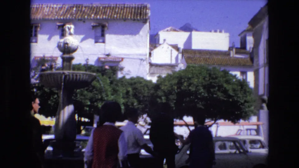 1972 Spain plaza view with a central stone fountain, whitewashed buildings, shaded trees, pedestrians, and classic compact cars along the street.