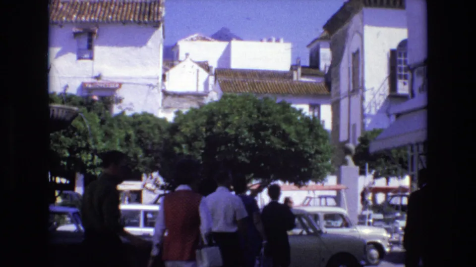 1972 Spain town-square frame with several pedestrians crossing in shade, whitewashed buildings, leafy trees, and compact cars parked along the curb.