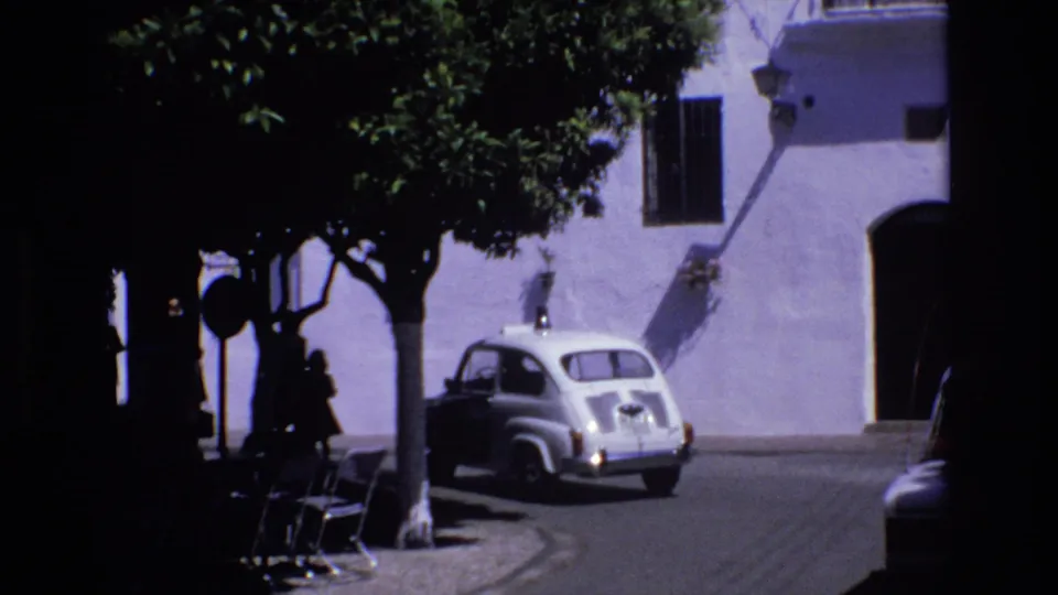 1972 Spain street-corner view showing a compact light-colored car by a whitewashed building, with trees, benches, deep shade, and a curving roadway.