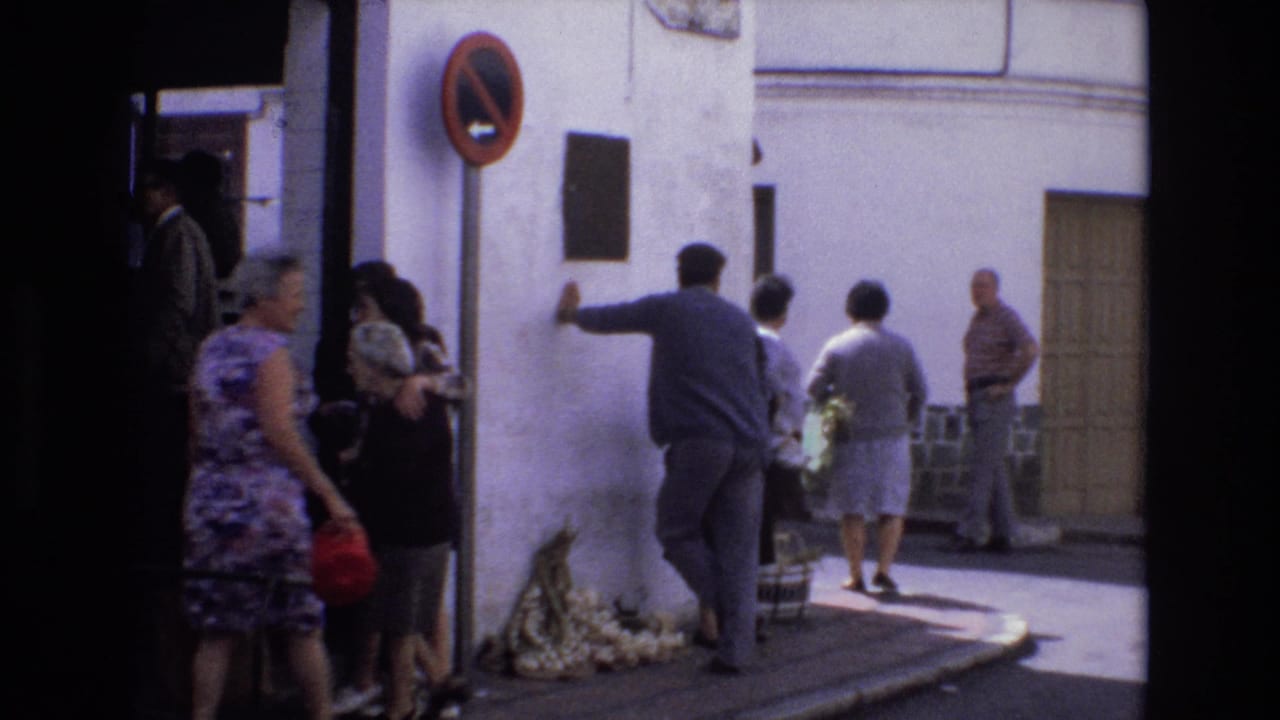 In 1972 Spain, adults stand and walk beside a whitewashed street corner as a man reaches toward the wall near a dark plaque and a no-parking sign.