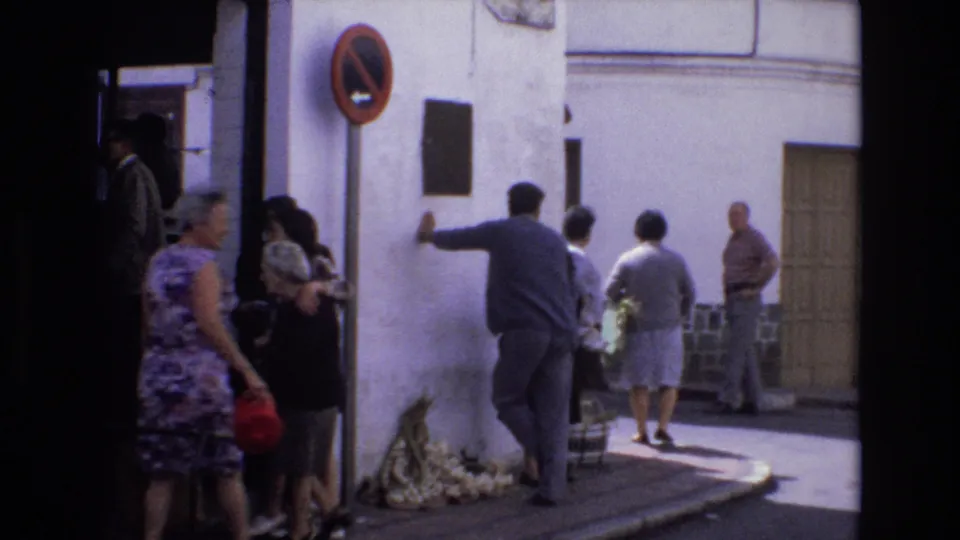 In 1972 Spain, adults stand and walk beside a whitewashed street corner as a man reaches toward the wall near a dark plaque and a no-parking sign.