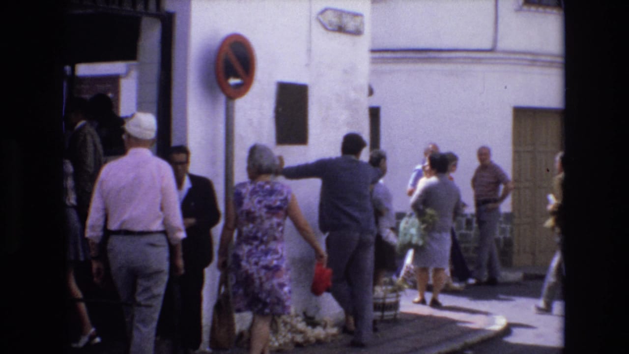 Pedestrians pass a white street corner while a man gestures toward the wall near a plaque.