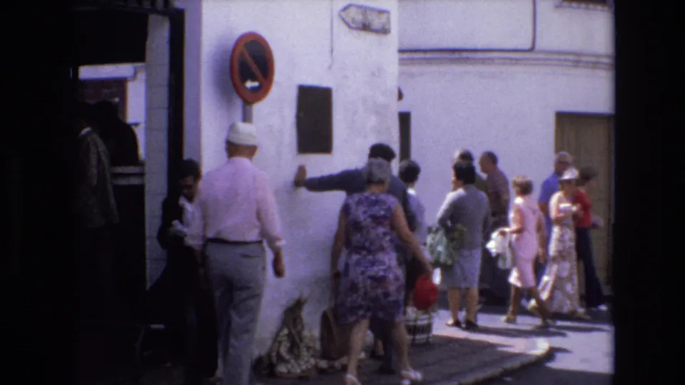 In 1972 Spain, a man remains near the wall while adults in dresses and light summer clothing pass through a narrow sunlit street near a plaque and traffic sign.