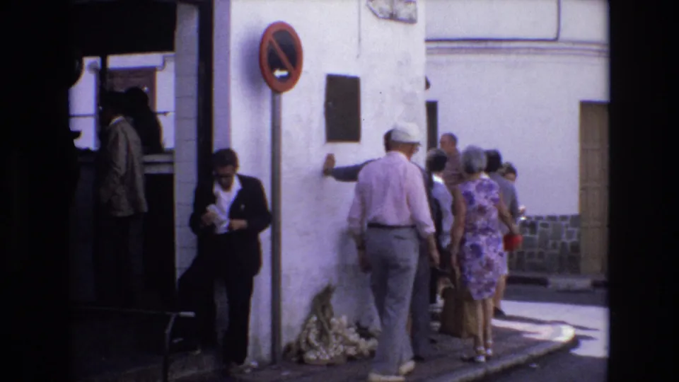 In 1972 Spain, adults continue past a white town street corner as the man near the wall and nearby pedestrians remain framed between deep shade and bright sunlight.