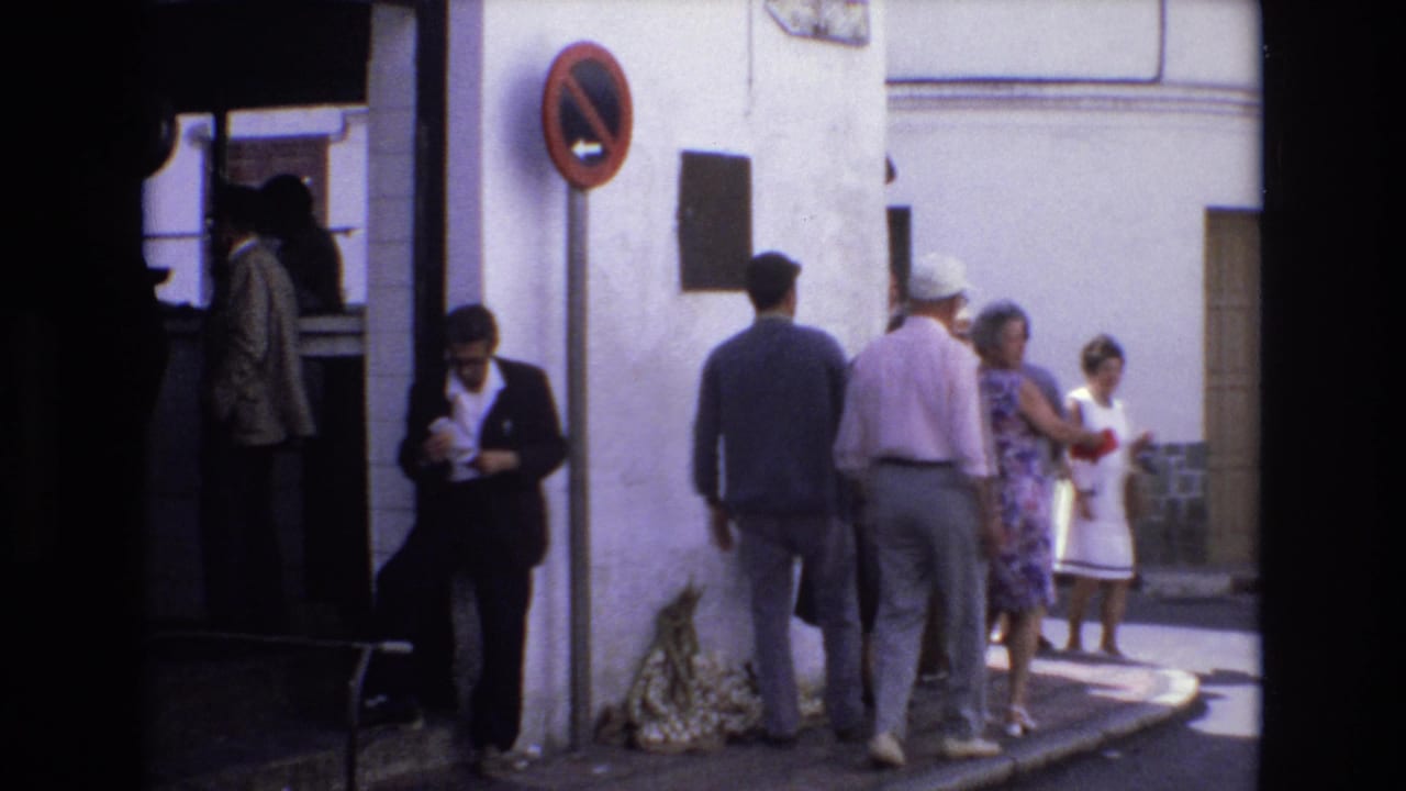 In Spain in 1972, adults stand and walk outside a whitewashed building with a red no-entry sign, doorway, and shaded sidewalk visible.