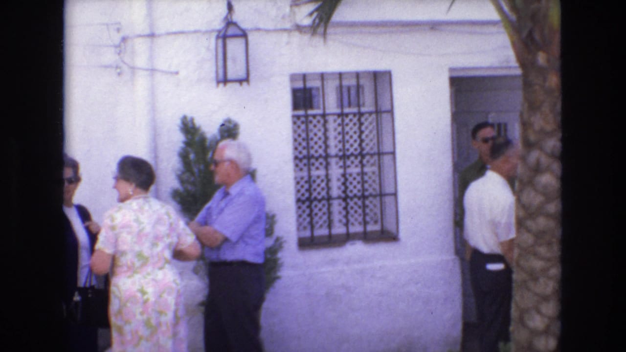 Adults stand near a barred window, wall lantern, doorway, and palm trunk outside a white building.