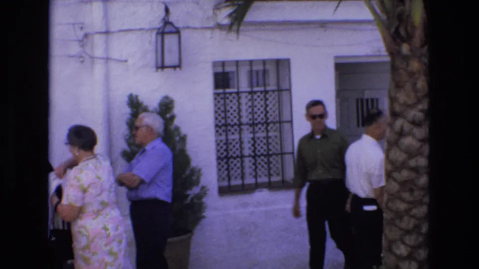 In Spain in 1972, adults in summer clothing stand and pass one another near a barred window, potted plant, doorway, and palm tree outside a white building.