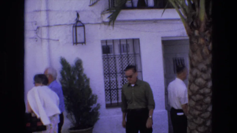 In Spain in 1972, adults move past a white building facade with a barred window, wall lantern, doorway, potted plant, and palm tree in view.