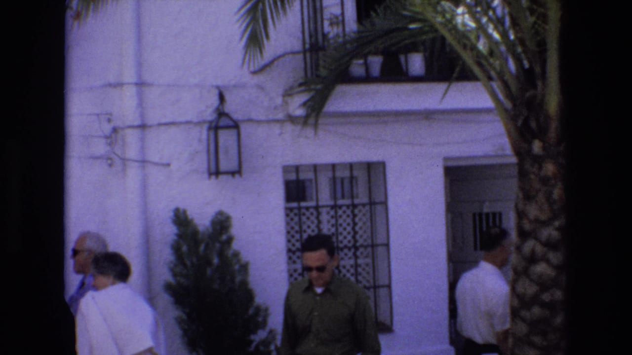1972 Spain home-movie frame showing several pedestrians near a white stucco facade with black window grilles, a wall lantern, shrubs, and a palm tree in the foreground.