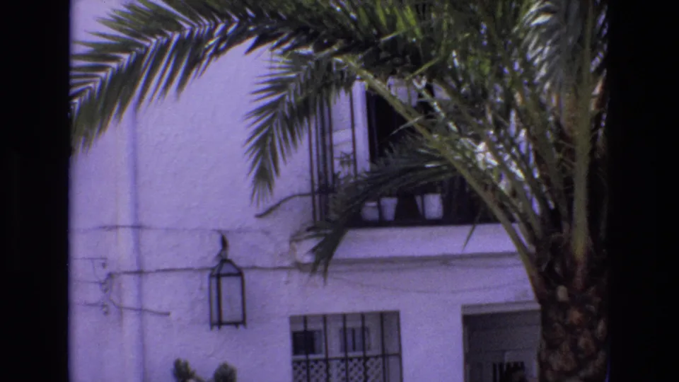 1972 Spain home-movie frame focused on the building exterior, with a palm tree in front, black window grille, wall lantern, and open doorway visible.