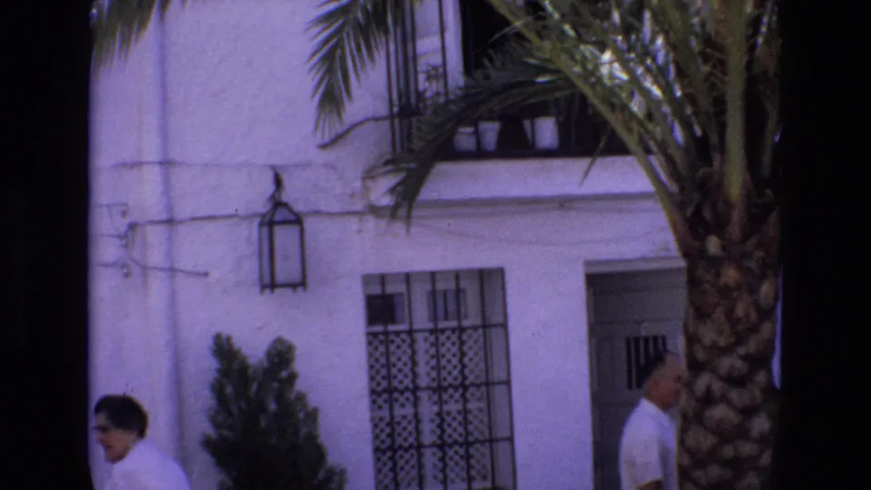 1972 Spain frame showing a man in a light shirt walking near a palm tree in front of a white stucco building with a black window grille and wall lantern.