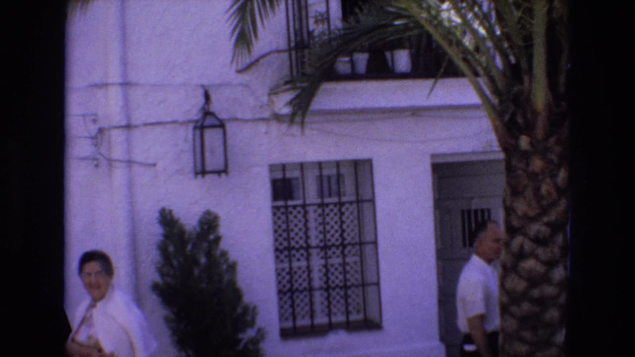 In 1972 Spain home-movie footage, a woman in white looks toward the camera outside a white stucco building with a man, palm tree, and wrought-iron window grille nearby.