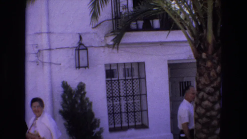 In 1972 Spain home-movie footage, a woman in white looks toward the camera outside a white stucco building with a man, palm tree, and wrought-iron window grille nearby.