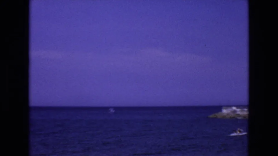 The camera pans across a calm Mediterranean seascape in 1972 Spain, showing saturated blue water, a distant boat, and a small rocky shoreline or island at frame right.