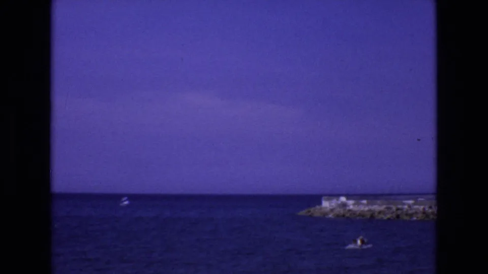 A final 1972 Spain seascape frame shows blue water, a rocky offshore point, a small distant boat, and a tiny figure or swimmer near the lower right.