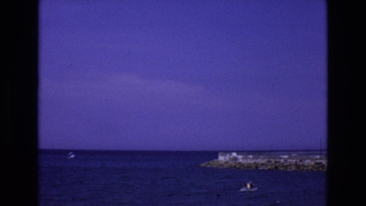 1972 Spain home-movie frame showing a rocky coastal pier, open sea, saturated sky, and a small boat with distant people near the lower edge.