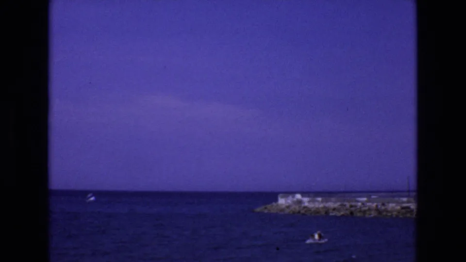1972 Spain home-movie frame showing a rocky coastal pier, open sea, saturated sky, and a small boat with distant people near the lower edge.