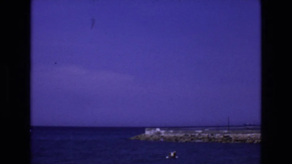 1972 Spain coastal view with a rocky pier at right, deep blue water, a small boat carrying distant figures, and a broad purple-blue sky.