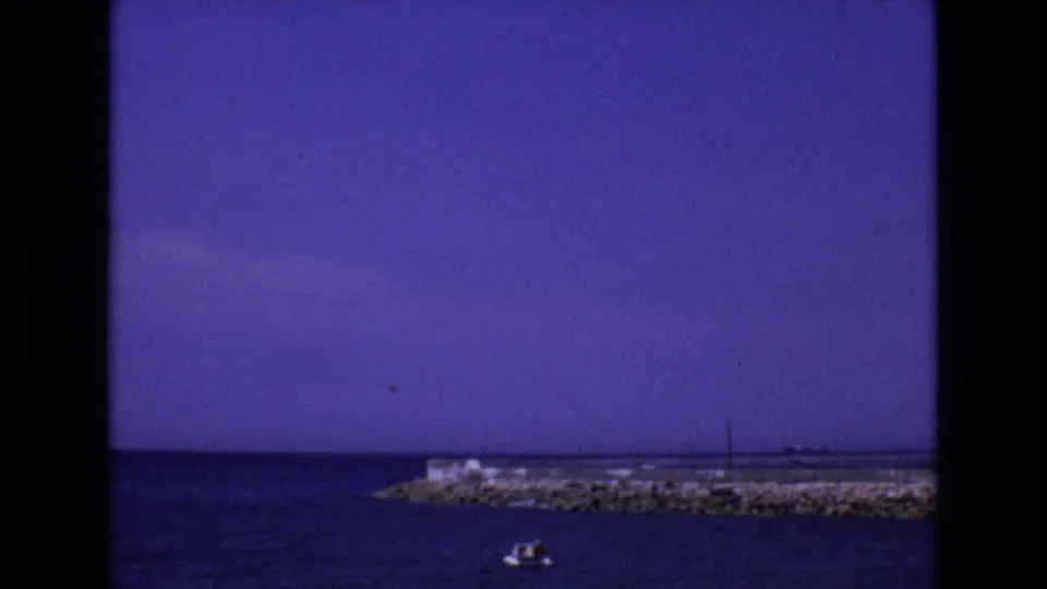 1972 Spain 8mm frame with a small boat in the foreground water, rocky pier across the background, and saturated sky above the coastline.