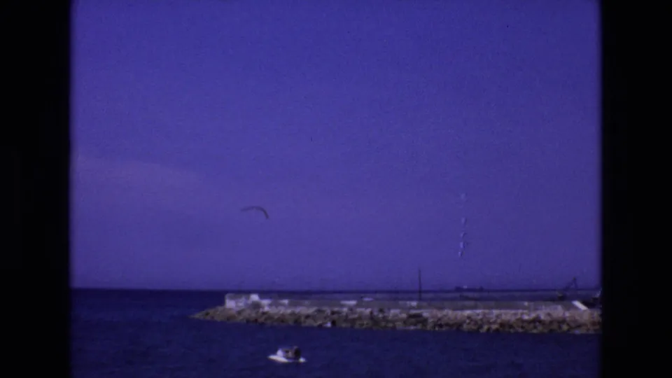 1972 Spain coastal home-movie frame showing a rocky breakwater, a small boat with distant people, open water, and faint vessel or mast shapes near the horizon.