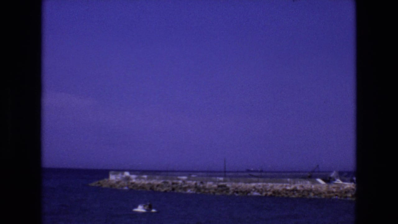 1972 Spain 8mm frame showing a small motorboat crossing calm coastal water in front of a rocky breakwater and distant shoreline structures.