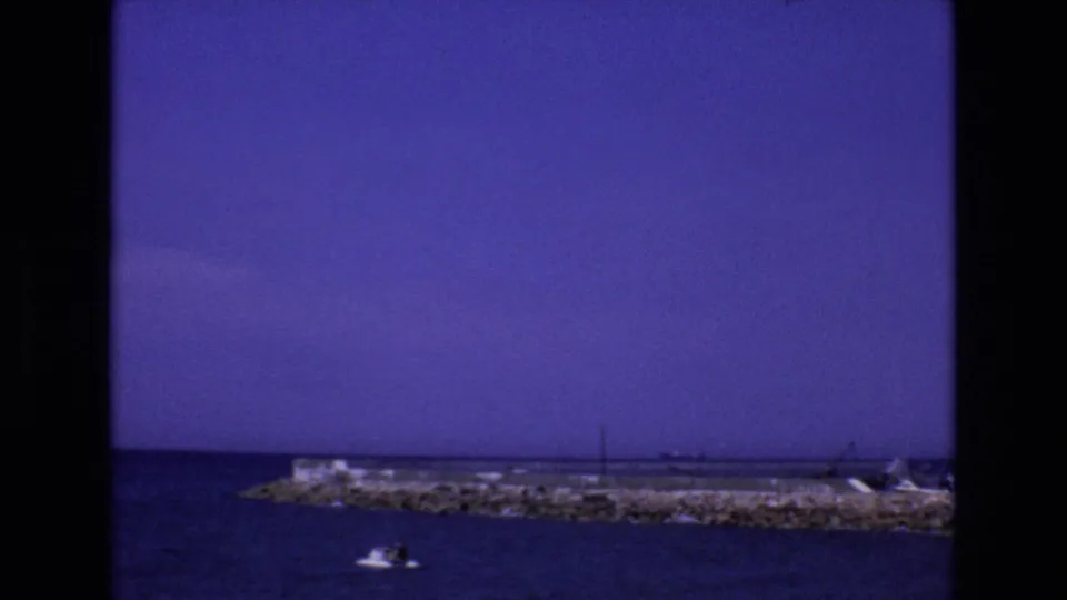 1972 Spain 8mm frame showing a small motorboat crossing calm coastal water in front of a rocky breakwater and distant shoreline structures.