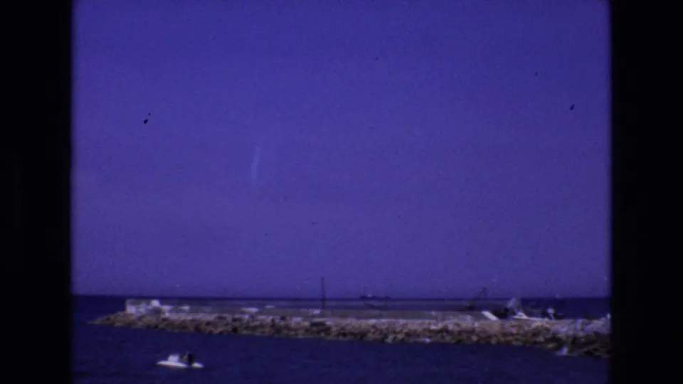 1972 Spain 8mm frame with a small motorboat in the foreground, a rocky breakwater across the middle distance, and faint shoreline structures beyond.