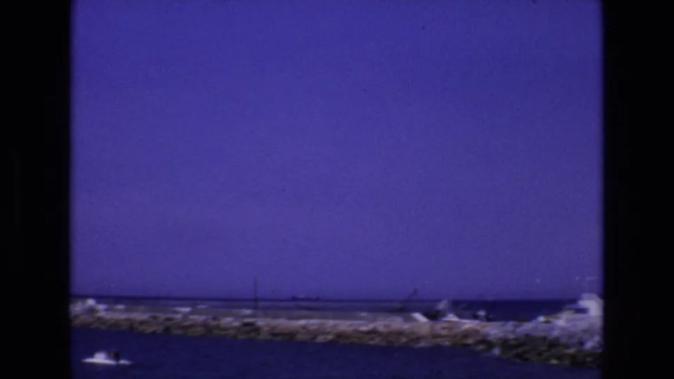 1972 Spain 8mm frame showing calm coastal water, a small boat near the lower edge, and a rocky breakwater stretching across the shoreline.