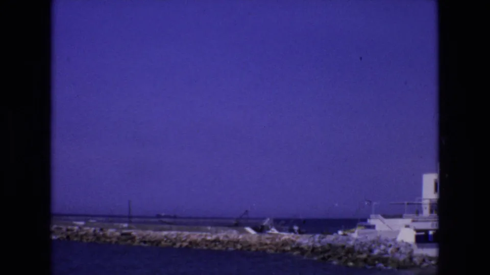 1972 Spain 8mm frame showing a rocky coastal breakwater, calm water, distant shoreline details, and part of a white waterfront building at the right edge.