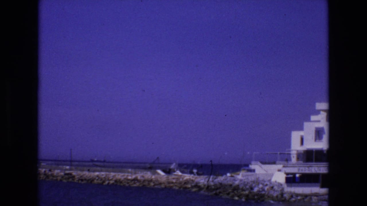 1972 Spain 8mm frame showing a white flat-roofed coastal building at the edge of a rocky breakwater, with ocean water and small boats nearby.