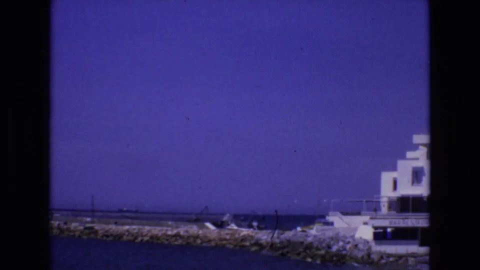 1972 Spain 8mm frame showing a white flat-roofed coastal building at the edge of a rocky breakwater, with ocean water and small boats nearby.