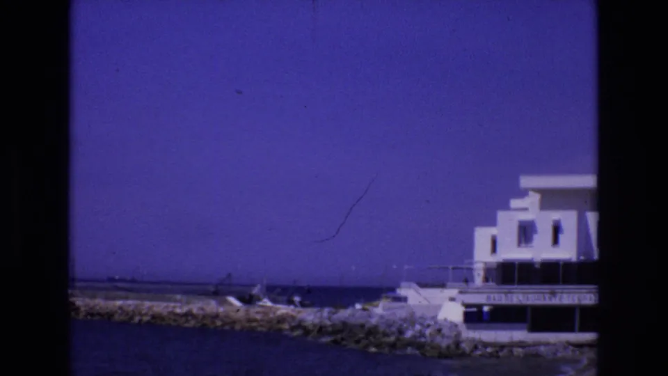 1972 Spain home-movie frame with a rocky breakwater, small boats near the waterline, and a white waterfront building partly visible at right.