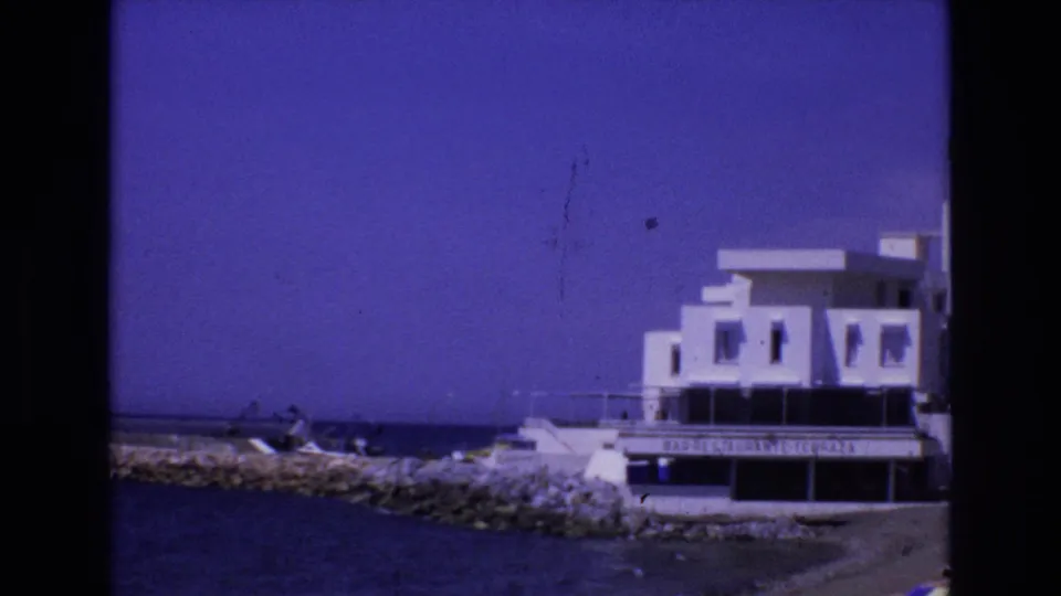 1972 Spain 8mm view of a white flat-roofed building facing the water, with rocky shoreline, small boats, and a low dock or seawall in front.