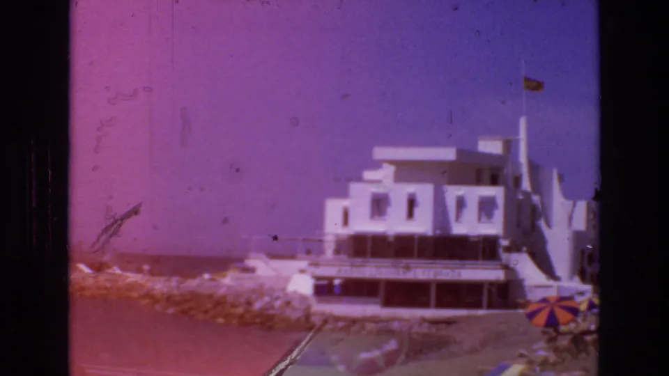 1972 Spain 8mm frame with a magenta color shift, showing the white coastal building, rocky shoreline, water, a small flag, and a striped beach umbrella.