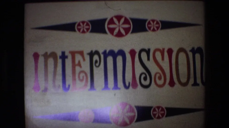 1972 Spain 8mm frame showing a projected intermission title card with multicolored serif lettering, black tapered bands, and red-and-white floral motifs.