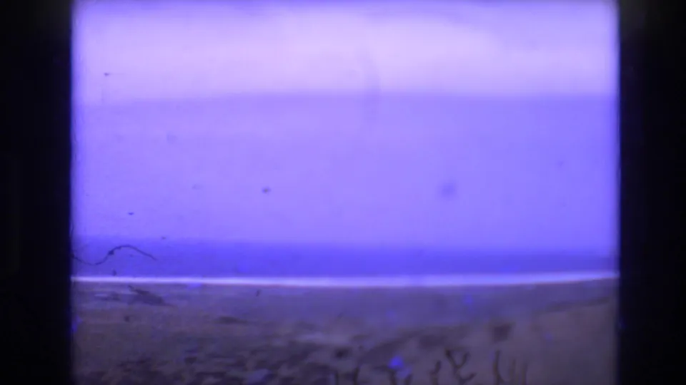 1972 Spain home-movie frame showing a blue-violet sky above a sandy beach and ocean horizon, with dark 8mm frame edges and visible dust.
