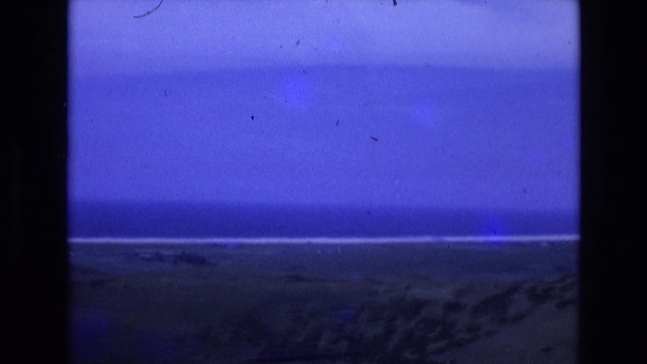 Ocean horizon and dim beach beneath a deep blue sky, with vertical black film border visible.